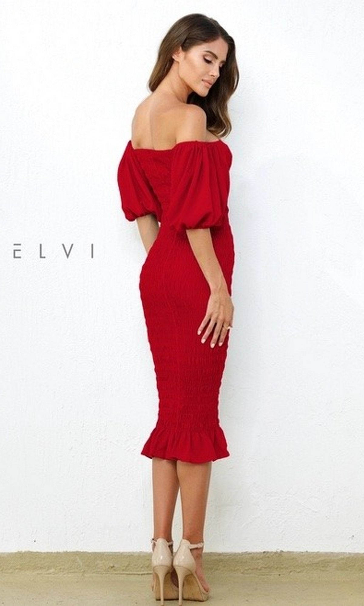 A woman stands against a white wall in the Skyla Knee-Length Puff-Sleeve Wedding-Guest Dress in red, featuring an off-the-shoulder design and ruffled hem, paired with nude heels—an elegant look for parties or weddings.